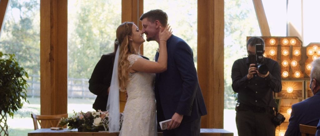 A still from wedding video with Bride and groom kissing during ceremony at the Mythe barn